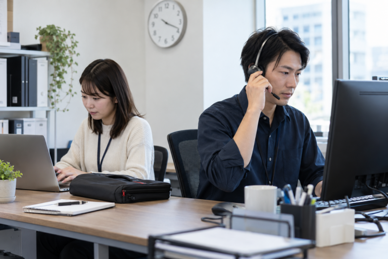 小さなPC設定サービスの事務所で、一人が相談電話を受け、もう一人がパソコンを確認する架空の成人二人
