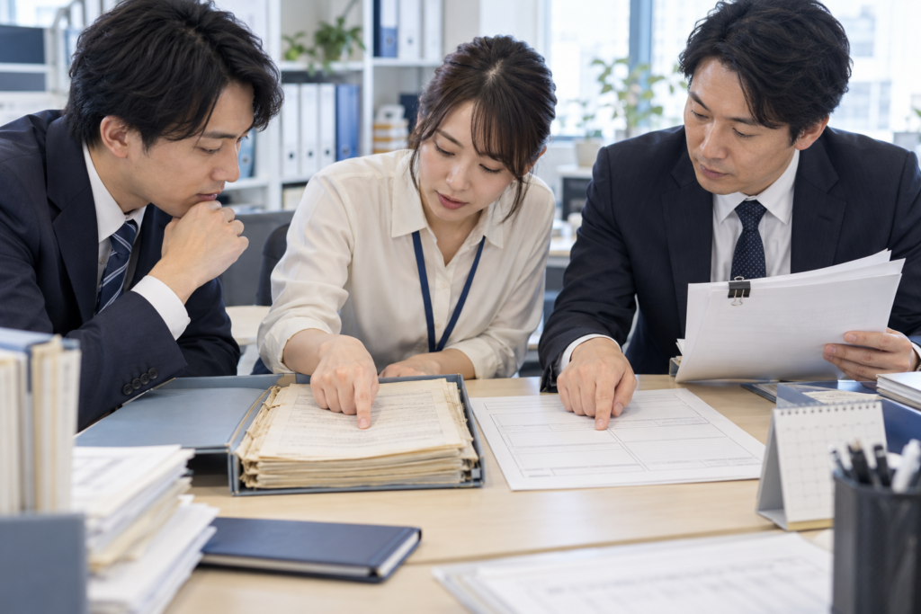 備品購入の案内を確かめるために、旧手順と現行手順を比べる架空の仕事の情景。